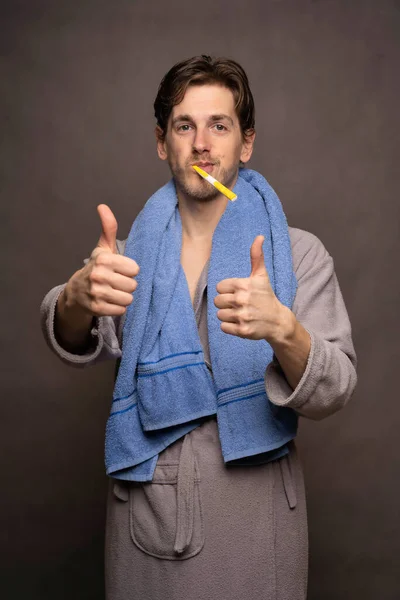 年轻英俊 瘦小的白人男子 棕色头发 灰色背景的灰色浴衣上用大拇指刷牙 — 图库照片
