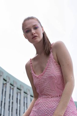 Beyaz, sarışın, at kuyruklu, pembe yazlık elbiseli, arkasında bina olan mavi gözlü genç bir kadın.