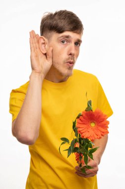 Genç, uzun boylu, zayıf, kahverengi saçlı, elinde turuncu çiçekle duymaya çalışan, beyaz arka planda izole edilmiş sarı gömlekli bir adam.