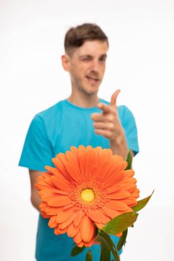 Genç, uzun boylu, zayıf, kahverengi saçlı, turuncu çiçek sunan, beyaz arka planda mavi gömlekli, yakışıklı beyaz bir adam.