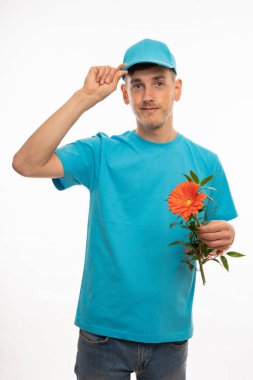 Genç yakışıklı, uzun boylu, zayıf, kahverengi saçlı, kepini düzelten, beyaz arka planda mavi gömlekli, turuncu çiçekli genç adam.