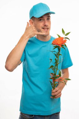 Genç, uzun boylu, zayıf, kahverengi saçlı, elinde turuncu çiçekle telefon işareti yapan beyaz arka planda izole edilmiş mavi gömlekli bir adam.