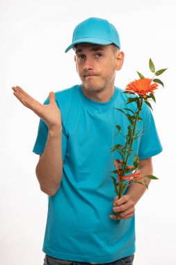 Genç yakışıklı, uzun boylu, zayıf, kahverengi saçlı, dikkatsiz görünen, elinde mavi gömlekli, mavi şapkalı, beyaz arka planda izole edilmiş bir turuncu çiçek tutan adam.