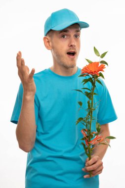 Genç yakışıklı, uzun boylu, zayıf, kahverengi saçlı beyaz adam elinde mavi gömlekli, mavi şapkalı, beyaz arka planda izole bir turuncu çiçek tutan bir şeyi açıklıyor.