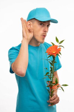 Genç, uzun boylu, zayıf, kahverengi saçlı, turuncu çiçekli, mavi gömlekli, beyaz arka planda yalnız bir adam duymaya çalışıyor.