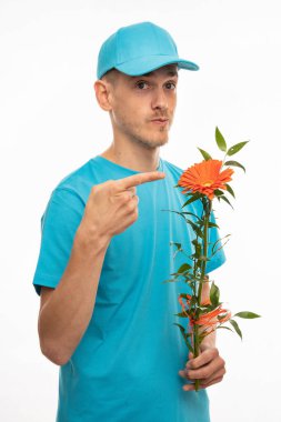 Genç, uzun boylu, zayıf, kahverengi saçlı, turuncu çiçekleri işaret eden, beyaz arka planda mavi gömlekli, yakışıklı, beyaz bir adam.