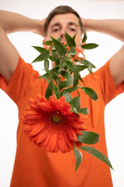 Genç, uzun boylu, zayıf, kahverengi saçlı, ağzı turuncu çiçekli, başı turuncu tişörtlü beyaz bir adam.