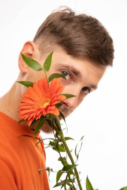 Genç, uzun boylu, zayıf, kahverengi saçlı, ağzı turuncu çiçekle kaplı, beyaz arka planda izole edilmiş turuncu gömlekli bir adam.