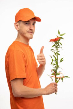 Yakışıklı, uzun boylu, zayıf, kahverengi saçlı, elinde turuncu çiçekle beyaz arka planda turuncu gömlekli genç bir adam.