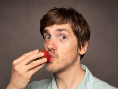 Genç yakışıklı, uzun boylu, zayıf, kahverengi saçlı, gri arka planda açık mavi gömlekli bir çileği öpen beyaz adam.