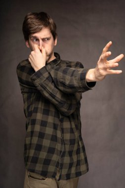 Genç yakışıklı, uzun boylu, zayıf, kahverengi saçlı, burnunu tutarak gri arka planda flanel gömleği olan beyaz bir adam.