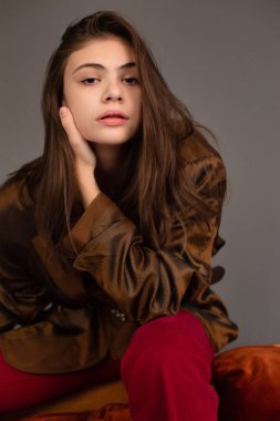 Uzun siyah saçlı, kahverengi gözlü, arkasında koyu gri duvar olan genç bir kadın.