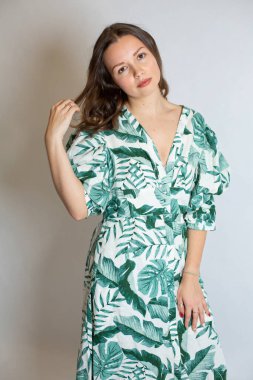 Çiçek desenli elbiseli çekici genç Avrupalı kadın saçlarına dokunurken gri renkte izole edilmiş.