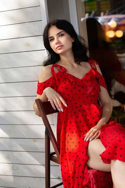 椅子に座って若いです黒髪の女性とともに白い肌で赤いドレスでカフェの近くに外 — ストック写真
