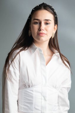 Siyah saçlı, beyaz, uzun kollu bluzlu, gri renkli, gülümseyen genç bir kadının portresi.