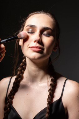 Makyaj portresi, koyu gri üzerine izole edilmiş gözleri olan iki örgülü genç Avrupalı kadını gülümsetiyor.