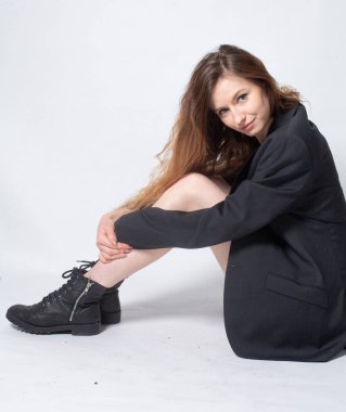 Siyah ceketli, beyaz, genç Avrupalı bir kadın, açık gri üzerinde, yerde tek başına oturuyor.