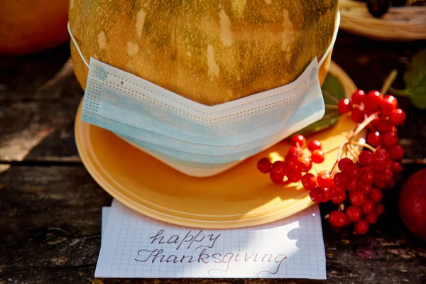 Pumpkin in mask and inscription Happy Thanksgiving. Autumn rustic concept with fruits, vegetables and medical mask with trendy shadows. Thanksgiving quarantine due to coronavirus concept.