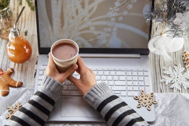 Kadın elleri karton bir bardak kakao tutuyor. Şenlikli ev ofisi: dizüstü bilgisayar, zencefilli çörek ve süslemeli Noel ağacı. Uzak çalışma kavramı, web sitesi ya da aile kavramı, kilitlenme nedeniyle arkadaşlar