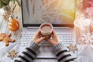 Kadın elinde bir fincan karton kahve tutuyor. Şenlikli ev ofisi: dizüstü bilgisayar, zencefilli kek ve son moda dekorasyonlu Noel ağacı. Kilitlenme kavramı yüzünden uzak çalışma, giysi ya da aile, arkadaşlar