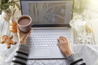 Şenlikli ev ofisi: dizüstü bilgisayar, zencefilli çörek ve süslemeli Noel ağacı. Kadın elleri karton bir bardak kakao tutuyor. Uzak çalışma kavramı, web sitesi ya da aile kavramı, kilitlenme nedeniyle arkadaşlar