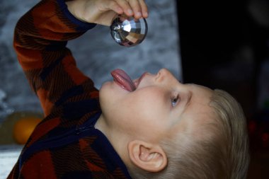 Komik neşeli çocuk disko topunu yalıyor. Çocuk Noel süslemeleri arasında Noel ağacı disko topuyla oynuyor. Yeni yıl arifesi için şenlikli atmosferik ev. Estetik bir ev. Mutlu erkek bebek