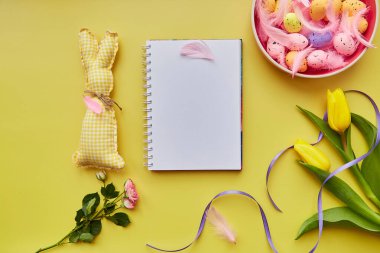 Bayramlık Paskalya not defteri gibi. Renkli yumurtalı pembe kase, pembe tüyler, el yapımı şirin tavşan ve çiçekler. Mutlu Paskalyalar. Sarı arkaplanda kartpostal modeli