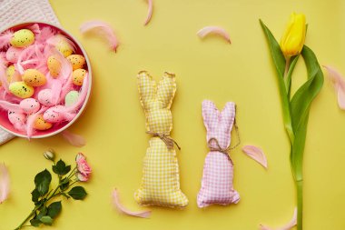 Bayramlık Paskalya kartı konsepti. Renkli yumurtalı pembe kase, pembe tüyler, sarı lale ve el yapımı sevimli tavşanlar. Yukarıdan görüntüle.