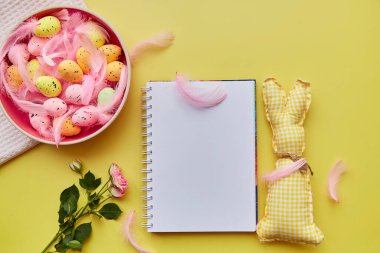Paskalya not defteri gibi. Renkli yumurtalı pembe kase, pembe tüyler, pembe gül ve el yapımı sevimli sarı tavşan oyuncağı. Mutlu Paskalyalar. Üst Manzara. Sarı arkaplanda kartpostal modeli