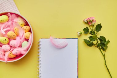 Paskalya not defteri. Renkli yumurtalı pembe kase, pembe tüyler ve pembe gül. Mutlu Paskalyalar. Üst Manzara. Sarı arkaplanda kartpostal modeli