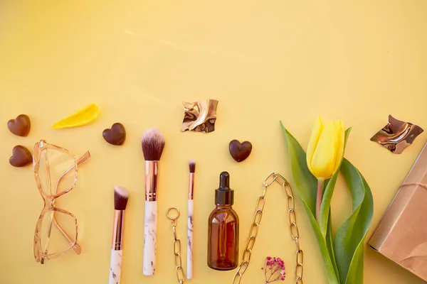 Trendy cosmetics and accessories on pastel yellow background with chocolate hearts and yellow tulips. Fashion glasses, makeup brushes, serum dropper, chain, earrings and gift box. Beauty card concept.