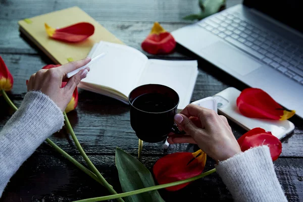 Girl holds cup of tea and pen near notepad and laptop. Online work, listening to music by headphones, learn language in app, online meeting, learning at home office with tulips. Taking webinar lesson.