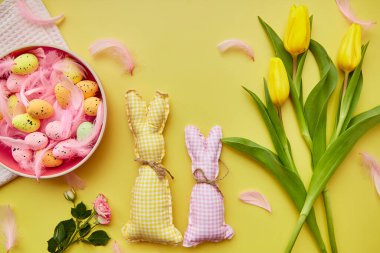 Bahar bayramı paskalya kartı konsepti. Renkli yumurtalı pembe kase, pembe tüyler, sarı laleler ve el yapımı sevimli tavşanlar.