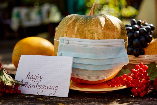 Pumpkin in mask and inscription Happy Thanksgiving. Autumn rustic concept with fruits, vegetables and medical mask with trendy shadows. Thanksgiving quarantine due to coronavirus concept.