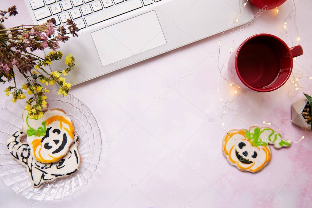 Cozy home office in Halloween : laptop and red cup, homemade cookies in shape of cute pumpkins. Atmospheric working atmosphere with garlands. Copy space.