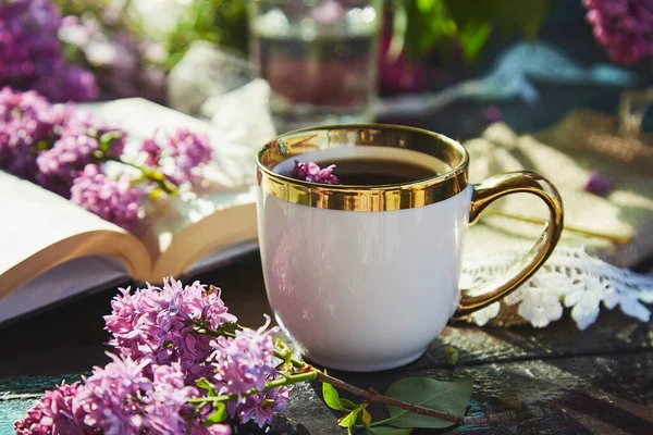 Romantic sunny garden, relax and leisure with cup of coffee. Mock up white cup and book surrounded lilac flowers. Cup of coffee, lilac flowers and book. Hard shadows. Summer days, aesthetic garden.