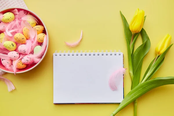 Festive notepad mock up. Pink bowl with colorful eggs, pink feathers and yellow tulips. Happy Easter concept. Post card mock up on yellow background