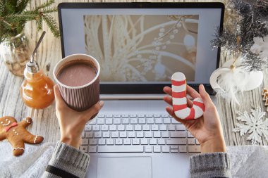 Kadın elleri karton bir fincan kakao ve zencefilli ekmek tutuyor. Şenlikli ev ofisi, dizüstü bilgisayar maketi, süslü Noel ağacı. Uzaktan çalışma, Webinar izlemek, çalışmak ya da aileyi aramak, arkadaşlar. Boşluğu kopyala