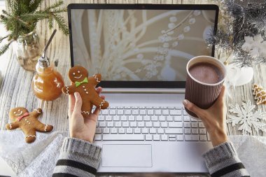 Kadın elleri karton bir fincan kakao ve zencefilli ekmek tutuyor. Şenlikli ev ofisi, dizüstü bilgisayar maketi, süslü Noel ağacı. Uzaktan çalışma, Webinar izlemek, çalışmak ya da aileyi aramak, arkadaşlar. Boşluğu kopyala