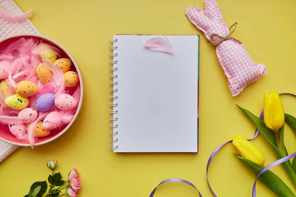Easter mock up notepad. Pink bowl with colorful eggs, pink feathers, handmade cute bunny and flowers. Happy Easter concept. Post card mock up on yellow background