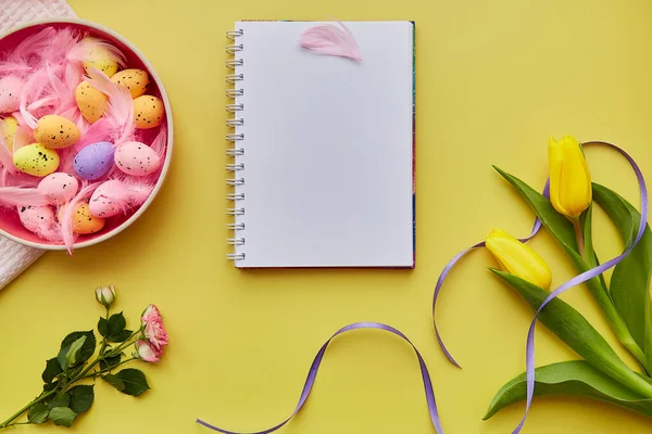 Easter notepad mock up. Colorful eggs in pink bowl, pink feathers and flowers. Happy Easter concept. Top view. Post card mock up on yellow background