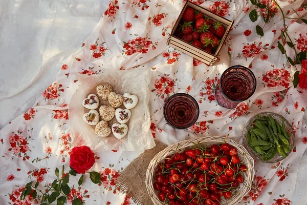 Pink summer picnic in the backyard. Flat lay tablecloth, box of marshmallows, strawberries, cherry, peas and glasses of wine among pink roses in the park. Romantic evening, appreciation of the moment