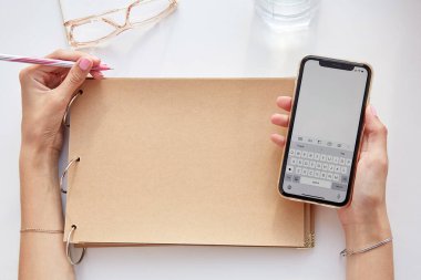 Woman writes by her left hand in a vintage crafting album while holding a phone. Glasess and glass of water. Conceptual photo of lefthanded day. Mock up screen phone. Copy space