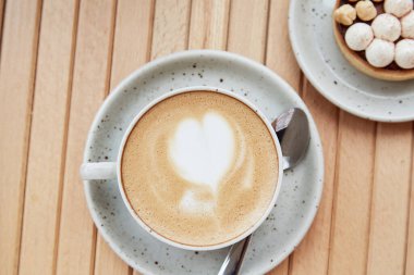 Heart shaped foam of cappuccino in white ceramic cup and hazelnut tart on wooden table at the terrace. Aesthetic breakfast top view. Flat lay food.