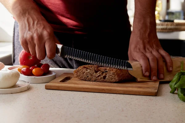 Preparing a dinner. Man is cutting a French fresh grain baguette in the kitchen. Basil, tomatoes and mozzarella on the table