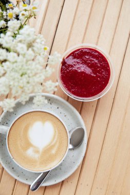 Cappuccino in a white ceramic cup with heart and raspberry jam dessert in a disposable cup - a snack, light breakfast on the cafe terrace. Flat lay food. Vertical photo.