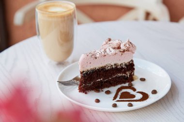 Fruit chocolate cake and a glass of latte on wooden table outdoor. Atmospheric summer breakfast. Pink flowers decorations in the cafe terrace.