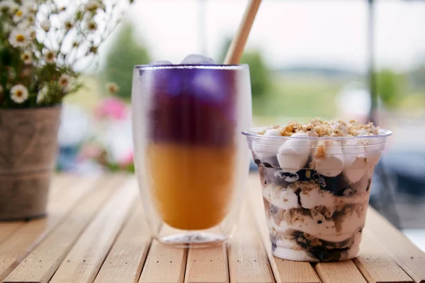 Vegan portion of nuts dessert in a disposable cup. Purple and yellow cocktail with straw reed - anchan blue tea with antioxidants and nootropics near chamomile flowers outside.