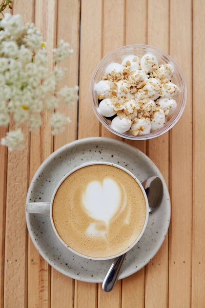 Ceramic white cup of cappuccino with heart, vegan portion of nuts dessert in a disposable cup - appetizer, light breakfast on cafe terrace with blurred foreground of chamomile flowers. Vertical photo.