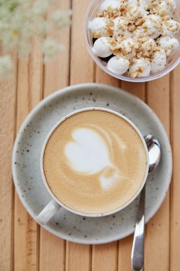 White cup of cappuccino with foam heart, vegan portion of nuts dessert in a disposable cup, light breakfast or coffee break with blurred foreground of chamomile flowers outside